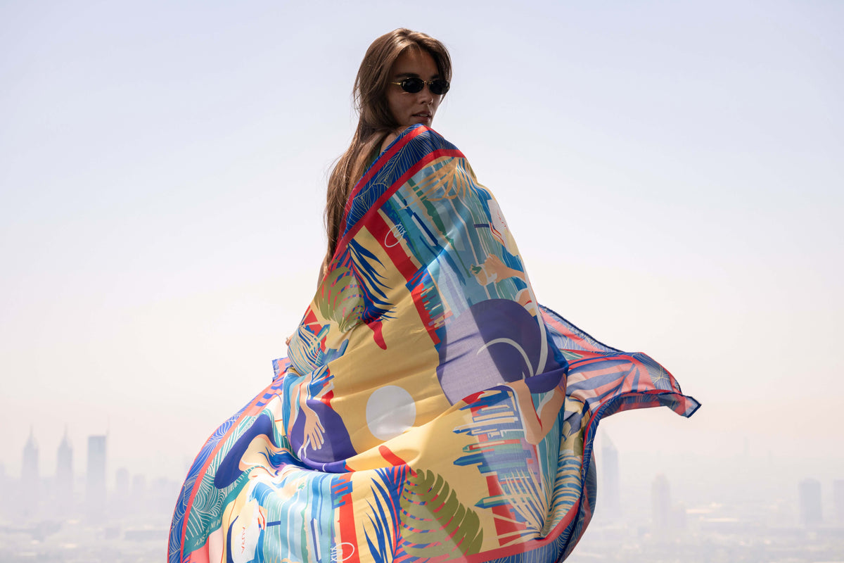 Aura Skypool Boutique campaign photo: a model holding a printed silk scarf with the Dubai skyline behind her